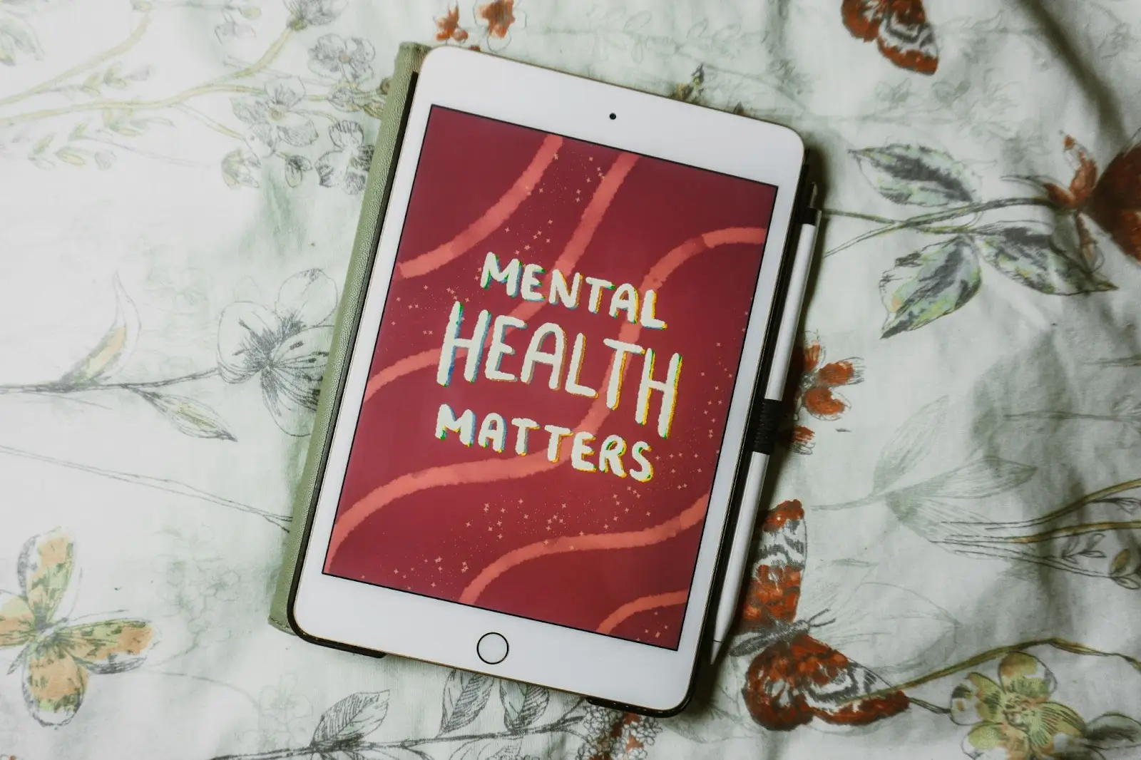 A tablet lying on a floral-patterned bedspread, displaying a red screen with the message “Mental Health Matters” in colorful, hand-drawn letters.