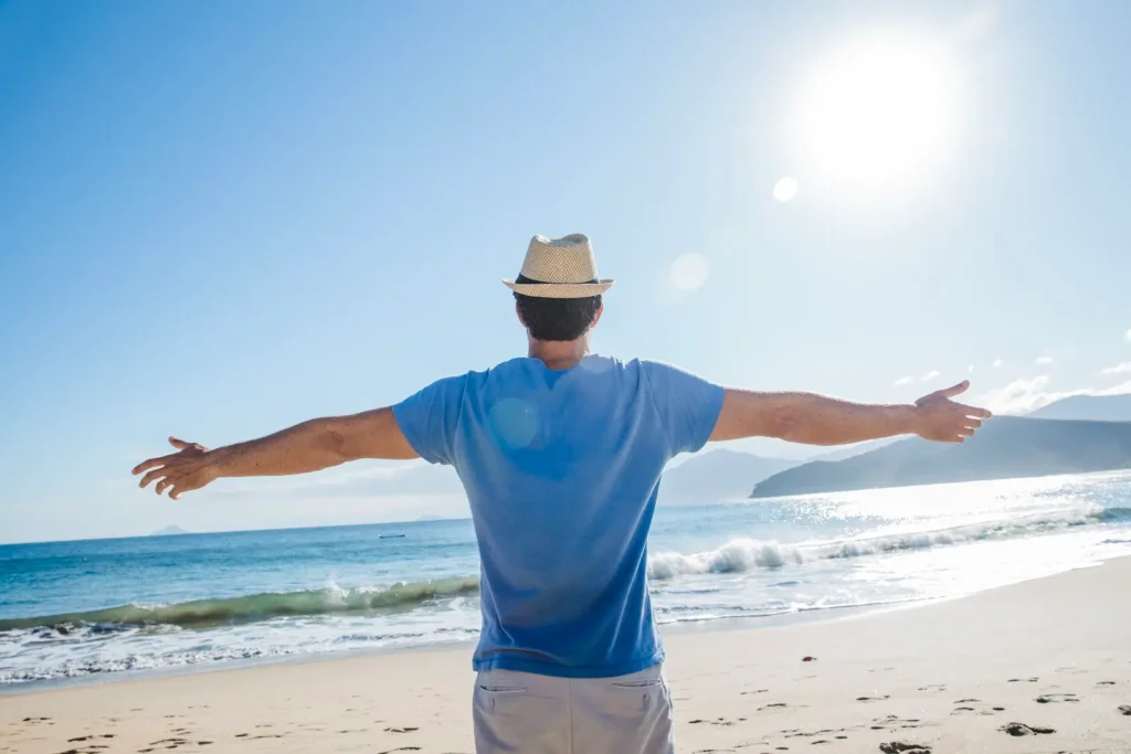 Man wearing a straw hat and blue t-shirt stands on a sunny beach with arms outstretched, facing the ocean and enjoying the scenic view of waves and distant mountains.