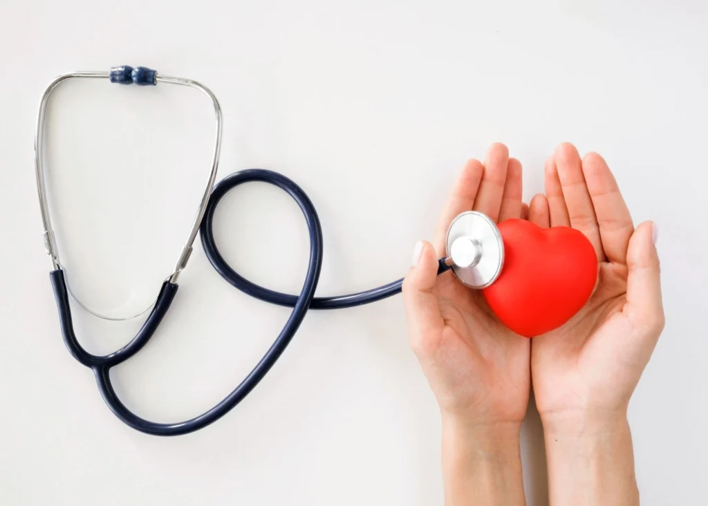 Hands holding a red heart model with a stethoscope on it.