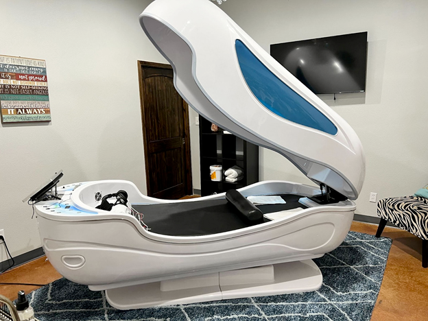 An open ozone sauna pod in a treatment room, with a blue-tinted window in its raised lid.